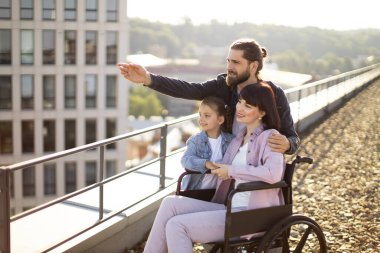 Modern binanın çatısında duran ve manzaranın tadını çıkaran beyaz bir ailenin yan görüntüsü. Tekerlekli sandalyedeki genç kadın anne aile desteğiyle yaralandıktan sonra rehabilitasyona giriyor..