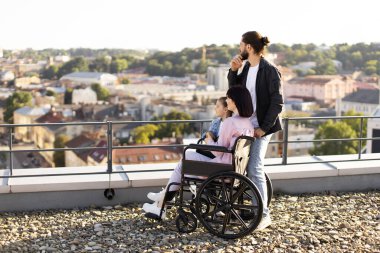 Tekerlekli sandalyedeki genç kadın anne aile desteğiyle yaralandıktan sonra rehabilitasyona giriyor. Modern binanın çatısında duran ve manzaranın tadını çıkaran beyaz bir ailenin yan görüntüsü..