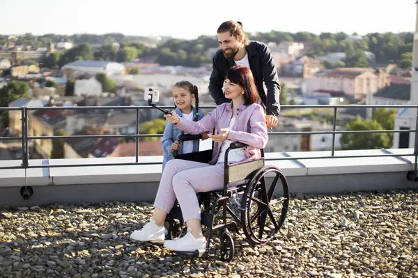 Tekerlekli sandalyedeki mutlu anne sevgi dolu bir kız ve kocanın bakımıyla çevrili. Genç bir aile evin çatısında iyi vakit geçiriyor, selfie çubuğu ve cep telefonu kullanarak fotoğraf çekiyor..