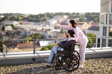 Genç beyaz anne, küçük tatlı kız ve tekerlekli sandalyede boncuklu baba modern evin çatısında gün batımını izliyor. Boş zaman, destek ve maluliyet kavramı.