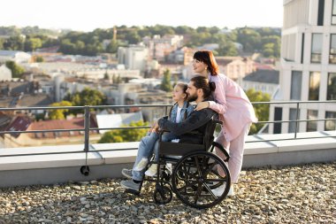 Genç beyaz anne, küçük tatlı kız ve tekerlekli sandalyede boncuklu baba modern evin çatısında gün batımını izliyor. Boş zaman, destek ve maluliyet kavramı.