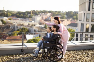 Boş zaman, destek ve maluliyet kavramı. Genç beyaz anne, küçük tatlı kız ve tekerlekli sandalyede boncuklu baba modern evin çatısında gün batımını seyrediyor..