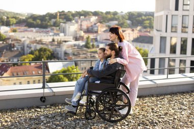 Boş zaman, destek ve maluliyet kavramı. Genç beyaz anne, küçük tatlı kız ve tekerlekli sandalyede boncuklu baba modern evin çatısında gün batımını seyrediyor..