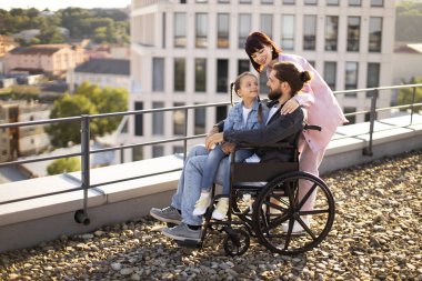 Modern binanın çatısında duran ve manzaranın tadını çıkaran beyaz bir ailenin yan görüntüsü. Tekerlekli sandalyedeki genç sakallı erkek baba, aile desteğiyle yaralandıktan sonra rehabilitasyona gidiyor..