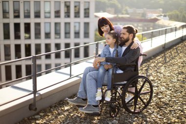 Modern binanın çatısında duran ve manzaranın tadını çıkaran beyaz bir ailenin yan görüntüsü. Tekerlekli sandalyedeki genç sakallı erkek baba, aile desteğiyle yaralandıktan sonra rehabilitasyona gidiyor..