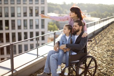 Tekerlekli sandalyedeki genç sakallı erkek baba aile desteğinde yaralandıktan sonra rehabilitasyona giriyor. Modern binanın çatısında duran ve manzaranın tadını çıkaran beyaz bir ailenin yan görüntüsü..