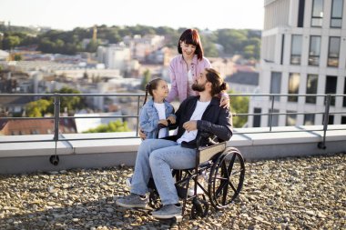 Tekerlekli sandalyedeki genç adam modern evin terasında ailesiyle eğleniyor. Anne ve kızı, babalarına temiz havada boş zaman geçirirken ciddi yaralanma tedavisi uyguluyor..