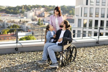 Anne ve kızı, açık havada boş zaman geçirirken ciddi yaralanma tedavisinde babalarına destek oluyor. Tekerlekli sandalyedeki genç adam modern evin terasında ailesiyle eğleniyor..