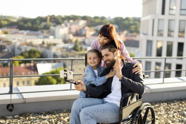Tekerlekli sandalyedeki gülümseyen adam çatıdaki manzaralı genç kadın ve kızla selfie çekiyor. Aile bağları ve neşeli anlar.