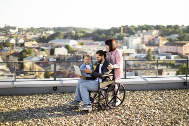 Mutlu aile çatıda dizüstü bilgisayarın keyfini çıkarıyor. Tekerlekli sandalyedeki adam, kadın ve kız şehir manzarasıyla kaynaşıyor..