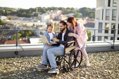 Mutlu aile çatıda birlikte vakit geçiriyor. Tekerlekli sandalyede genç kızı ve yanında destekleyici bir kadınla dizüstü bilgisayar kullanan bir adam..