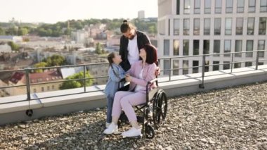 Özürlü genç bir ailenin portresi modern evlerle şehir planına karşı. Genç bir adam ve küçük bir kız tekerlekli sandalyede tedavi gören anneyle ilgileniyorlar..