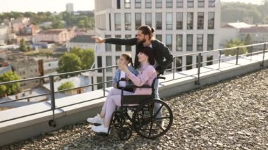 Modern binanın çatısında duran ve manzaranın tadını çıkaran beyaz bir ailenin yan görüntüsü. Tekerlekli sandalyedeki genç kadın anne aile desteğiyle yaralandıktan sonra rehabilitasyona giriyor..