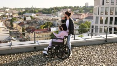 Tekerlekli sandalyedeki genç kadın anne aile desteğiyle yaralandıktan sonra rehabilitasyona giriyor. Modern binanın çatısında duran ve manzaranın tadını çıkaran beyaz bir ailenin yan görüntüsü..