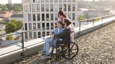 Boş zaman, destek ve maluliyet kavramı. Genç beyaz anne, küçük tatlı kız ve tekerlekli sandalyede boncuklu baba modern evin çatısında gün batımını seyrediyor..
