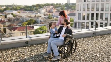 Tekerlekli sandalyedeki genç adam modern evin terasında ailesiyle eğleniyor. Anne ve kızı, babalarına temiz havada boş zaman geçirirken ciddi yaralanma tedavisi uyguluyor..