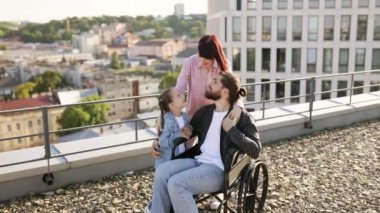 Tekerlekli sandalyedeki genç adam modern evin terasında ailesiyle eğleniyor. Anne ve kızı, babalarına temiz havada boş zaman geçirirken ciddi yaralanma tedavisi uyguluyor..