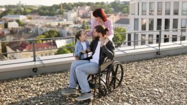Özürlü genç bir ailenin portresi modern evlerle şehir planına karşı. Genç bir kadın ve küçük bir kız, tekerlekli sandalyede tedavi gören babasıyla ilgileniyorlar..