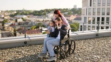 Tekerlekli sandalyede babasıyla mutlu bir aile çatıdaki şehir manzaralı bir terasta selfie çekiyor. Birlikte çekilen neşeli an.