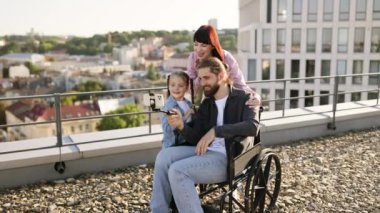Çocuk ve ebeveynlerin dışarıda selfie çektiği neşeli bir aile anı. Babam tekerlekli sandalye kullanıyor, mutluluğu ve birlikteliği ifade ediyor..