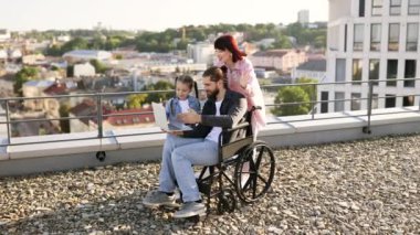 Mutlu bir aile, tekerlekli sandalyedeki bir adam da dahil olmak üzere çatıda birlikte dizüstü bilgisayar kullanarak kaliteli zaman geçiriyorlar. Arka planda manzara şehir görünümü.
