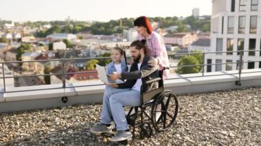 Mutlu aile çatıda birlikte vakit geçiriyor. Tekerlekli sandalyede genç kızı ve yanında destekleyici bir kadınla dizüstü bilgisayar kullanan bir adam..