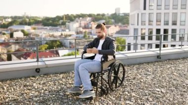 Tekerlekli sandalyedeki genç beyaz adam şehir çatısı üzerinde akıllı telefon kullanıyor, daktilo ediyor ya da tarıyor. Modern şehir yaşamında bağımsızlık, teknoloji ve ulaşılabilirlik.