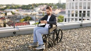 Tekerlekli sandalyedeki sakallı adam şehrin çatısında akıllı telefon kullanıyor, teknolojiyi ve olumluluğu kucaklıyor..