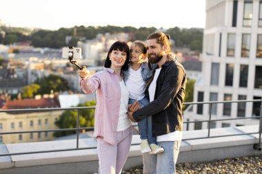 Üç kişilik mutlu bir aile, anne-baba ve kız, arka planda güzel bir şehir manzarasıyla çatıda selfie çekiyorlar..