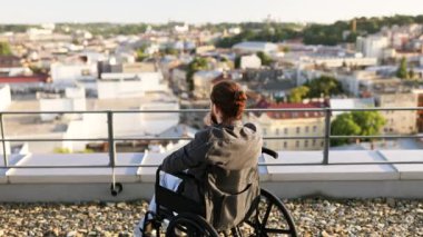 Tekerlekli sandalyedeki adam, kollarını kaldırmış, başarıyı kutlarken çatıdaki manzarayı izliyor..