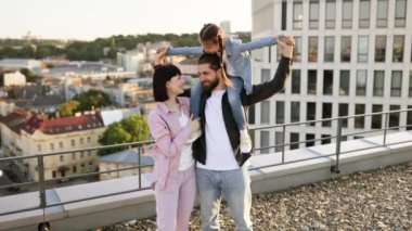 Üç kişilik beyaz bir aile çatıda eğleniyor. Kadın omuzlarında dururken, erkek bir kızı sırtında taşır..