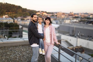 Mutlu aile çatıda birlikte vakit geçiriyor ve arka planda güzel bir şehir manzarası var. Ebeveynler ve kızları dışarıda gülüyor ve kaynaşıyorlar..