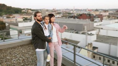 Genç bir aile çatıdan panoramik manzaranın keyfini çıkarıyor, gün batımında mutlu bir an, şehir manzarası arka planı.