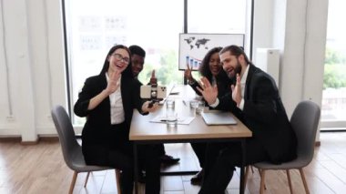 Modern ofiste toplantı sırasında dört kişilik iş ekibi selfie çekiyor. Resmi giyinmiş iş arkadaşları gülümsüyor, el hareketlerini gösteriyor ve takım ruhu yaratıyor..