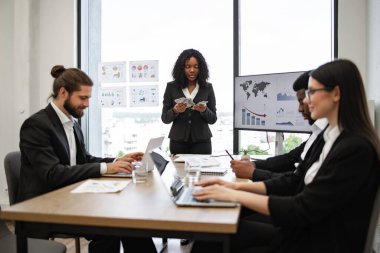 Afrikalı Amerikalı iş kadını iş toplantısında para sayıyor, ekip mali raporları tartışıyor, ekrandaki grafikler, duvardaki tablolar, modern ofis ortamı