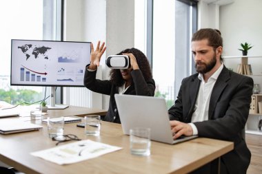 Ofis masasında resmi giyinmiş iş arkadaşları. Biri sanal gerçeklik kulaklığı kullanırken diğeri dizüstü bilgisayarla çalışıyor. Arkaplanda monitörde gösterilen grafik ve grafikler, modern ofis ayarları.