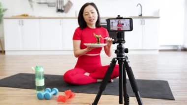 Spor kıyafetleri içinde bir kadın akıllı telefon kullanarak evde sağlıklı beslenme videosu çekiyor. Yoga minderinde sağlıklı salata, su şişesi ve egzersiz malzemeleriyle oturmak..