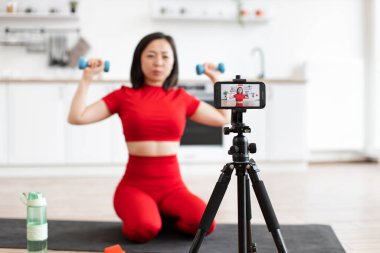 Kırmızı spor giyimli bir kadın akıllı telefon ve tripodu kullanarak evde fitness videosu çekiyor. O, halterleri tutuyor ve modern parlak mutfakta egzersiz yapıyor. Ev işi ve sağlıklı yaşam tarzı kavramı.