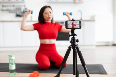 Kırmızı spor elbiseli bir kadın evde akıllı telefondan spor salonu videosu kaydederken dambıllarıyla egzersiz yapıyor. Sağlık, fitness ve çevrimiçi koçluk kavramlarını içerir.