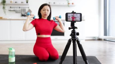 Kırmızı spor giyimli bir kadın akıllı telefon ve tripodu kullanarak evde fitness videosu çekiyor. O, halterleri tutuyor ve modern parlak mutfakta egzersiz yapıyor. Ev işi ve sağlıklı yaşam tarzı kavramı.