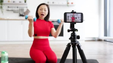 Kırmızı spor elbiseli bir kadın evde akıllı telefondan spor salonu videosu kaydederken dambıllarıyla egzersiz yapıyor. Sağlık, fitness ve çevrimiçi koçluk kavramlarını içerir.