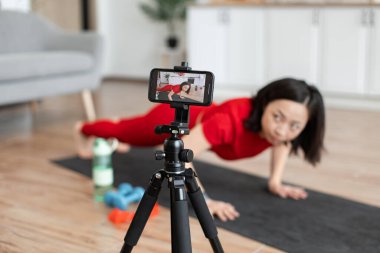 Kırmızı elbiseli kadın yoga minderinde şınav çekerken akıllı telefondaki fitness videosunu kaydediyor. Egzersiz ekipmanları arka planda görünür.