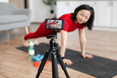 Kırmızı elbiseli bir kadın kendini akıllı telefondan kaydederken şınav çekiyor. Arka planda görünür haldeki dambıl ve su şişesi gibi egzersiz ekipmanları.