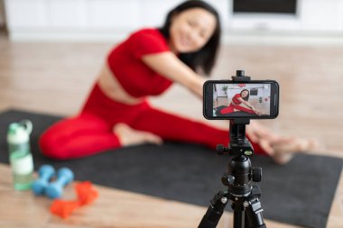 Kadın akıllı telefondan çekim yaparken evde yoga yapıyor. Kırmızı etek giyiyor ve yoga minderi ve halterler kullanıyor. Sanal uygunluk içeriği oluşturuluyor.