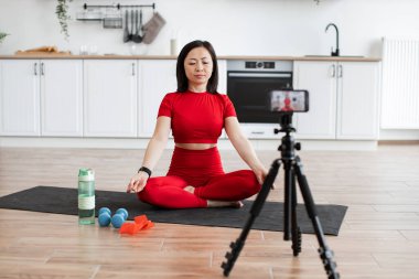 Kadın akıllı telefonuyla video kaydederken mutfakta yoga minderi üzerinde meditasyon yapıyor. Egzersiz aletleri ve bir şişe su görünür. Sakin ve odaklanmış atmosfer.