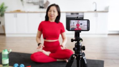 Bir kadın akıllı telefonuyla video kaydederken minder üzerinde yoga meditasyonu yapıyor. Zindelik ve rahatlama kavramı, temiz ve modern bir mutfak ortamında kaydedildi..