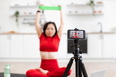 Kırmızı spor elbiseli bir kadın online egzersiz videosu çekerken direniş bandıyla geriniyor. Egzersiz modern bir ev ortamında, kameralar onun üzerinde yoğunlaşırken yapılır..