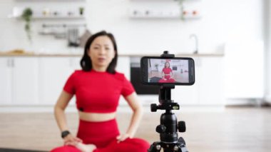 Spor kıyafetli bir kadın yoga minderi üzerinde dua pozisyonunda meditasyon yapıyor. Parlak mutfakta internetten yoga seansı kaydediyor. Evde spor yaparken etrafta spor aletleri var..