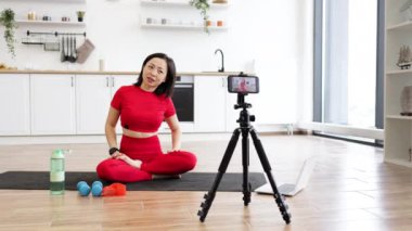 Kırmızı elbiseli bir kadın evde yoga yaparken online dersleri laptopta takip ediyor, modern mutfak ortamında yoga minderinde geriniyor.