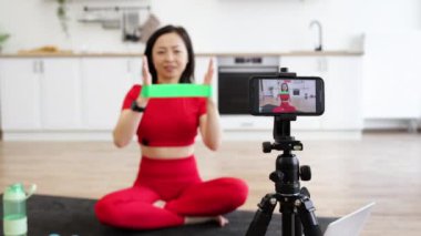 Kırmızı spor giysili bir kadın çevrimiçi fitness dersinde direniş bandıyla egzersiz yapıyor. Akıllı telefon, dizüstü bilgisayar, su şişesi ve dambıllarla çekiyoruz. Modern mutfak arka planı.